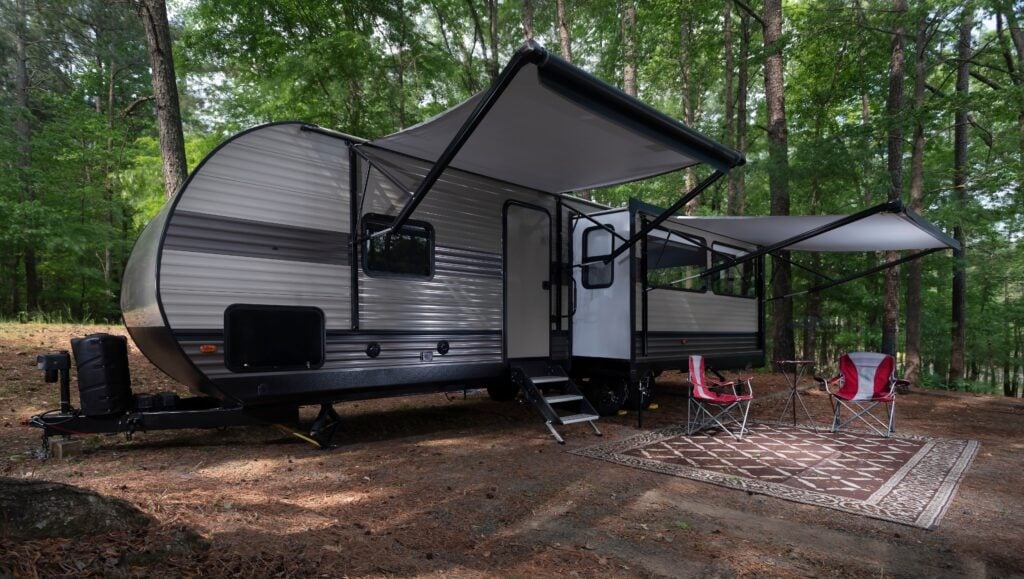Used RV set up in woods with 2 camp chairs outside it. Awnings are open.