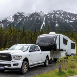 White Ram truck towing a Brinkley fifth-wheel RV on a scenic mountain road with snow-capped peaks and pine trees in the background, showcasing a luxury RV lifestyle surrounded by nature.