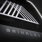 A black and white close-up photograph of the Brinkley RV logo, featuring a stylized triangular graphic design resembling abstract mountain peaks above the 'BRINKLEY' text, which appears in modern, spaced lettering. The image has a sleek, high-contrast aesthetic with metallic-looking text against a dark background.