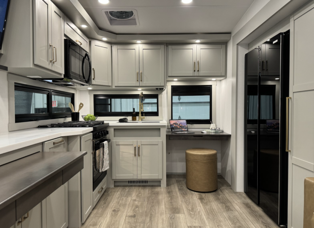 Modern kitchen and workspace area in the Brinkley Model Z 2680 RV, featuring sleek cabinetry, a built-in microwave, a spacious countertop with a stainless steel sink, a gas stove, and a compact desk setup with a round ottoman and a laptop.