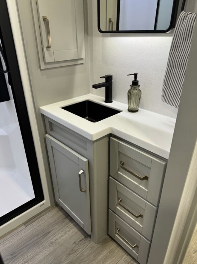 RV bathroom vanity in the Brinkley Model Z 2680, featuring a black sink with a matching modern faucet, a sleek white countertop, cabinetry with brushed gold handles, and a striped hand towel hanging nearby.