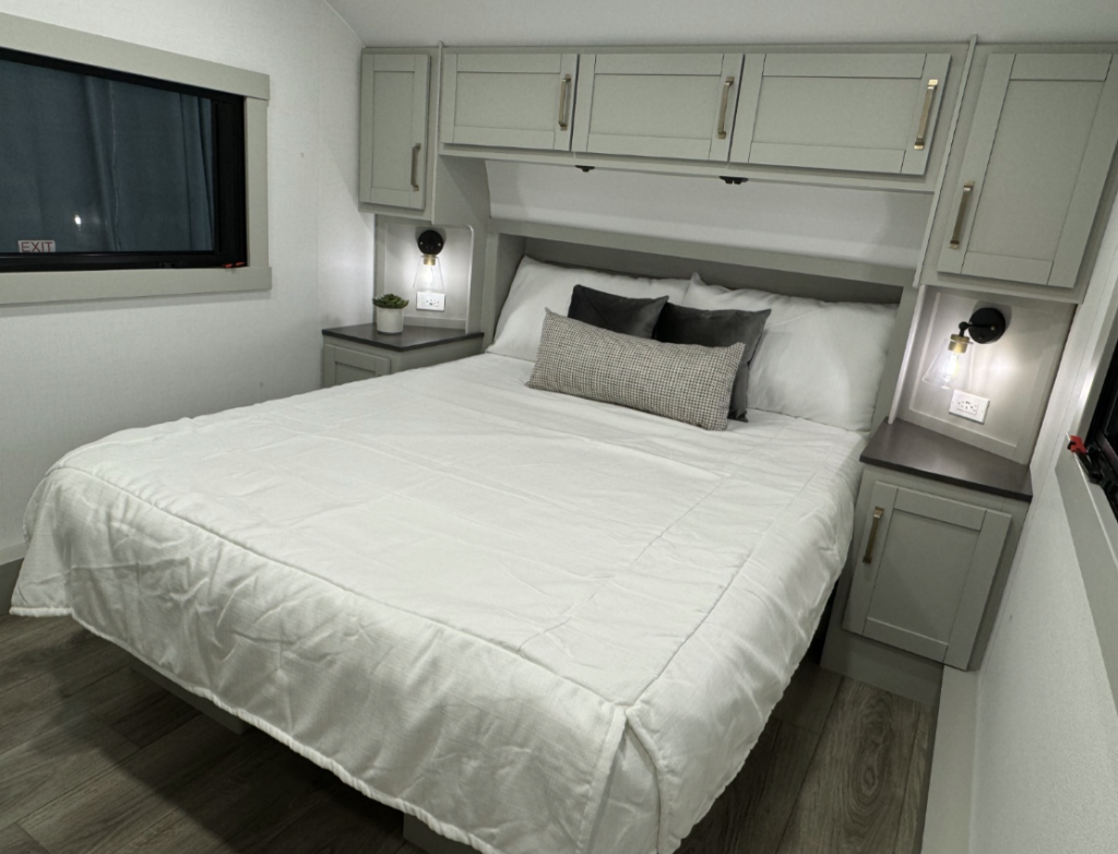 Bedroom in the Brinkley Model Z 2680 RV, featuring a neatly made bed with a white comforter and decorative pillows, flanked by nightstands with built-in cabinets and modern sconces, along with upper storage cabinets for additional organization.