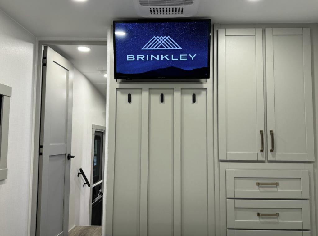 Interior view of the Brinkley Model Z 2680 RV, featuring a mounted flat-screen TV displaying the Brinkley logo, wall hooks for hanging items, and built-in cabinetry with drawers for organized storage, with a glimpse of a hallway leading to other areas.