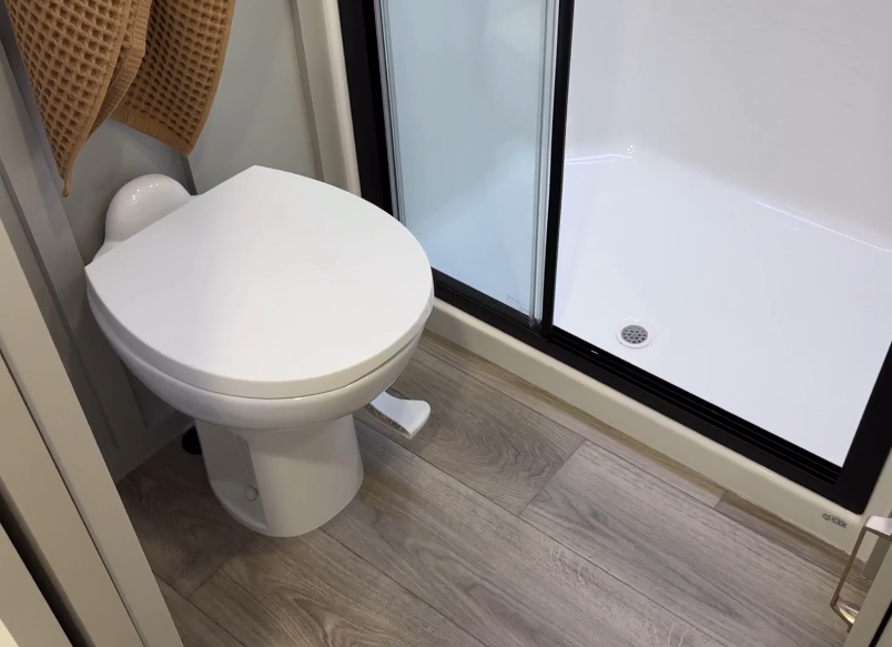 Bathroom in the Brinkley Model Z 2680 RV, featuring a sleek white toilet with a foot flush mechanism next to a spacious shower enclosure with a sliding glass door, set against stylish wood-look flooring.