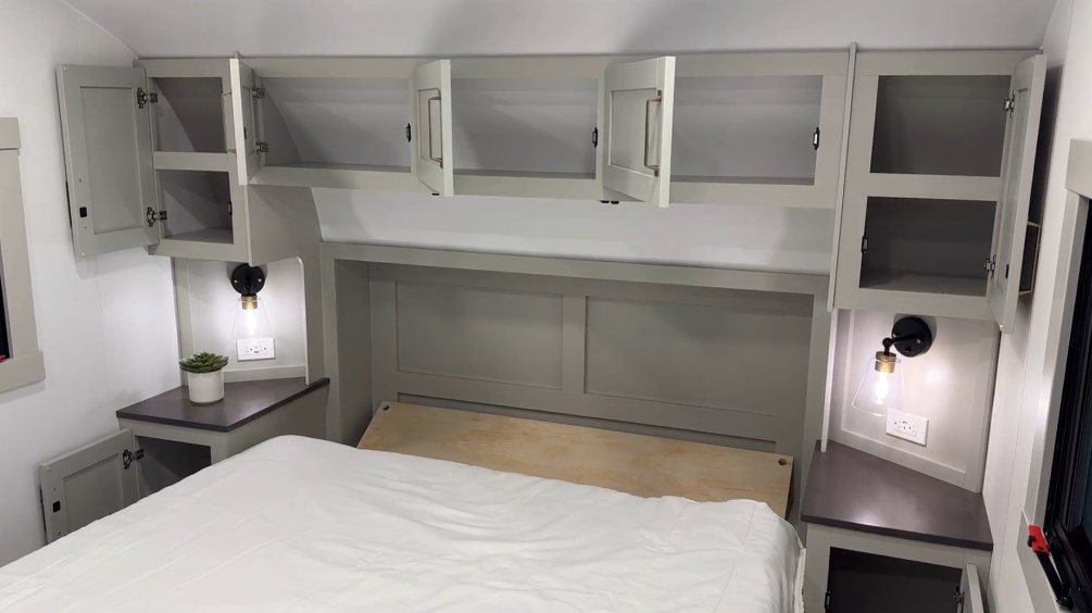 Bedroom in the Brinkley Model Z 2680 RV, featuring open overhead cabinets for storage, two bedside tables with built-in shelves, modern black sconces, and a partially made bed with a white comforter.