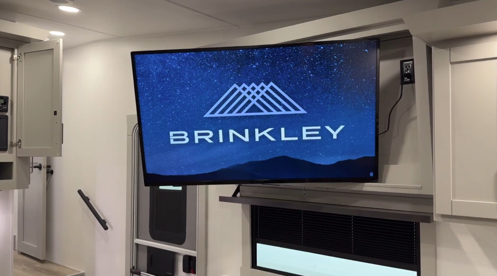 Mounted flat-screen television in the Brinkley Model Z 2680 RV, displaying the Brinkley logo over a starry night sky backdrop, positioned above modern cabinetry and next to a partially open storage cabinet.