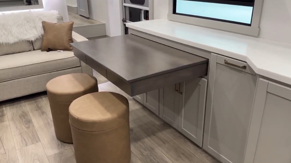 Pull-out dining table in the Brinkley Model Z 2680 RV, extended from the cabinetry with two round leather ottomans positioned underneath, creating a functional and stylish dining or workspace area in the living space.