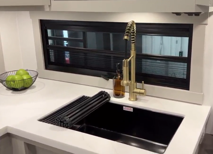 Modern RV kitchen sink in the Brinkley Model Z 2680, featuring a deep black basin, a luxurious gold pull-down faucet, and a roll-up drying rack, with a backdrop of a wide window and a bowl of green apples on the countertop.