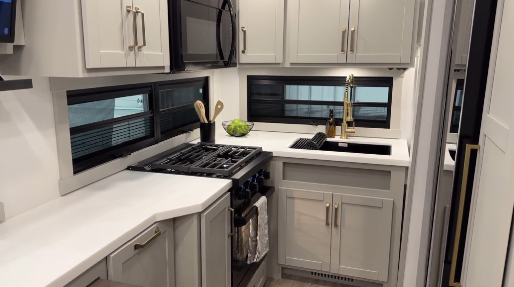 Corner kitchen in the Brinkley Model Z 2680 RV, featuring modern cabinetry, a built-in microwave, a gas stove with a four-burner cooktop, a deep stainless steel sink with a stylish faucet, and wide windows for natural light.