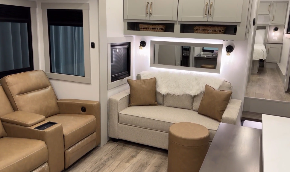 Interior of the Brinkley Model Z 2680 RV, showcasing a cozy living area with a beige reclining armchair, plush sofa adorned with decorative pillows and a soft throw, modern cabinetry, and a view into the adjoining bedroom.