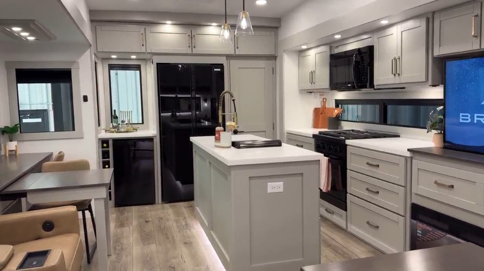 Spacious and modern RV kitchen featuring a central island with a white countertop and gold faucet, surrounded by light gray cabinetry with gold hardware. The fully equipped space includes a black stove, microwave, and refrigerator, accented by under-cabinet lighting. A wine rack and bar area add a touch of luxury, while pendant lights and wood-look flooring enhance the sleek and inviting design.