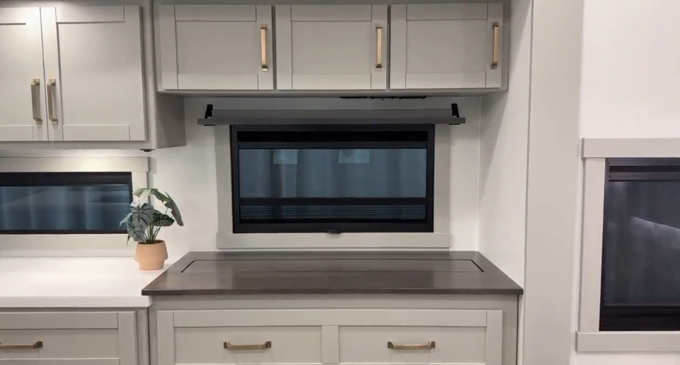 Contemporary RV kitchen space featuring light gray cabinetry with gold hardware, a dark wood countertop, and a compact window providing natural light. Upper cabinets offer ample storage, while a small potted plant adds a touch of greenery to the clean, modern design.