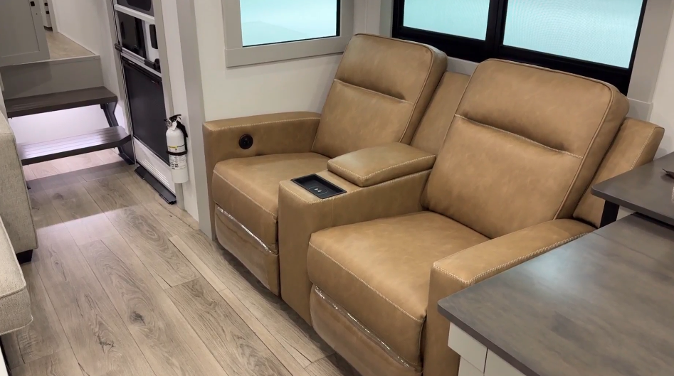 Contemporary RV seating area featuring a pair of tan leather recliners with built-in cup holders and a central armrest for added comfort. The recliners are positioned near large windows for natural light and overlook wood-look flooring, complementing the modern interior design. A compact kitchen setup is visible in the background, along with neatly integrated stairs leading to an upper section.