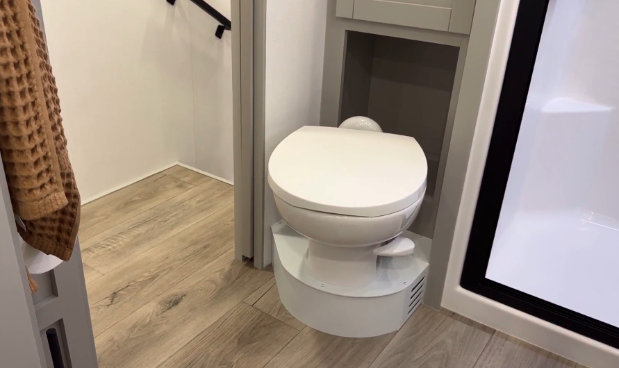 Compact RV bathroom featuring a modern white toilet with a raised base, surrounded by light gray cabinetry with open storage for added functionality. The design is complemented by wood-look flooring, a nearby glass shower enclosure, and warm decorative accents like a textured brown towel. The layout maximizes space while maintaining a sleek, clean aesthetic.