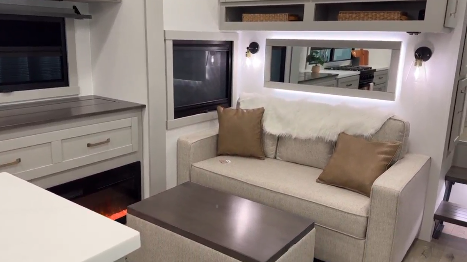 Modern RV interior featuring a cozy beige sofa with white faux fur and tan accent pillows, a dark wood coffee table, and an electric fireplace beneath a sleek entertainment setup. Illuminated shelving with baskets, contemporary wall sconces, and a clean white countertop enhance the minimalist, home-like ambiance.