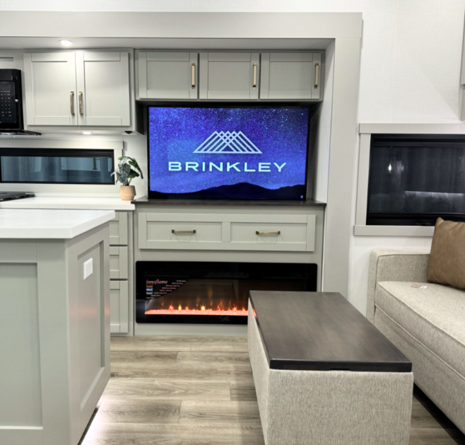 Modern RV interior showcasing a stylish entertainment center with a large TV displaying the 'Brinkley' logo, set above an electric fireplace with glowing flames. Surrounding light gray cabinetry with gold handles provides storage and a seamless design. Adjacent to the TV is a white countertop with a small plant for a fresh touch, while a beige sofa and coffee table complete the cozy, functional living space.