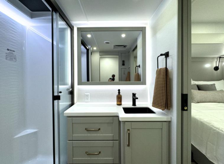 Modern RV bathroom featuring a sleek vanity with light gray cabinetry, gold hardware, and a white countertop with a black sink and faucet. An illuminated mirror adds brightness, while an amber soap dispenser and textured brown hand towel provide stylish accents. The design is complemented by a spacious glass-enclosed shower with bright white paneling, seamlessly connecting to the adjacent bedroom for a functional and cohesive space.