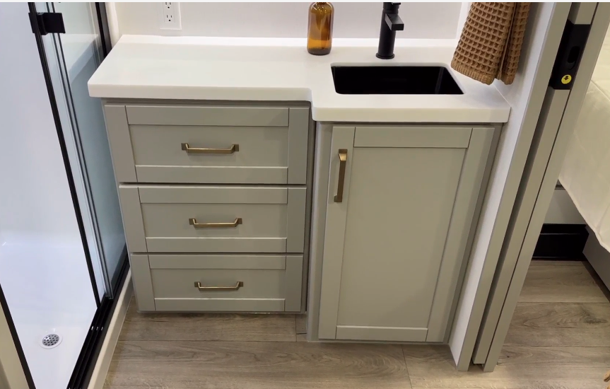 Compact RV bathroom vanity featuring light gray cabinetry with gold hardware, a white countertop, and a black sink with a matching modern faucet. Three drawers and a cabinet provide ample storage, while a soft brown hand towel and amber glass soap dispenser add warm, decorative accents. The wood-look flooring complements the clean and functional design, adjacent to a shower enclosure.