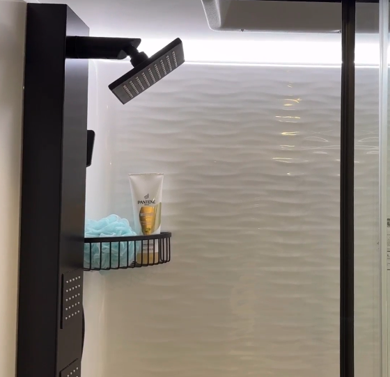 Modern RV shower with a sleek black rainfall showerhead and textured white wall paneling for a contemporary look. A black metal shelf holds shower essentials, including a blue loofah and shampoo, while recessed lighting enhances the bright and clean design. The glass enclosure adds a polished, spa-like touch.