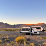 Fifth-wheel RV camper parked in scenic desert campsite at sunset, with red rock cliffs and mesa formations in background, yellow desert brush in foreground, under clear blue sky in Utah or Arizona landscape