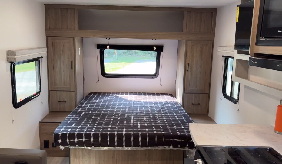 Modern RV bedroom featuring a cozy bed with a checkered bedspread, surrounded by light wood cabinets for ample storage, a large window for natural light, and functional side shelving for added convenience.