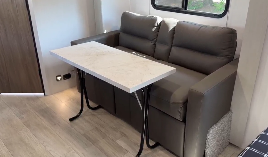 Versatile RV interior featuring a sleek gray loveseat paired with a functional, marble-style foldable dining table, set on light wood-style flooring in a modern, space-efficient design.