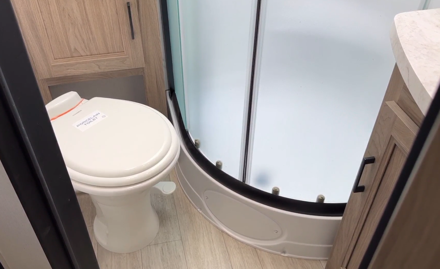 Modern RV bathroom featuring a porcelain toilet, a curved frosted glass shower enclosure, and light wood cabinetry, combining compact design with a clean, functional aesthetic.