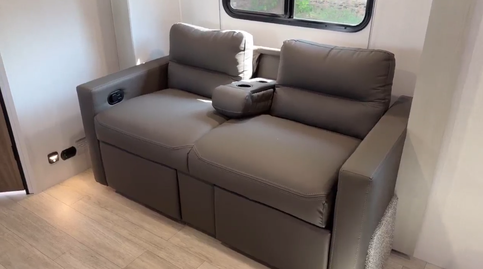 Modern RV loveseat with sleek gray upholstery, integrated cupholders, and storage drawers, set in a bright, minimalist RV interior with wood-style flooring and natural light from a window.