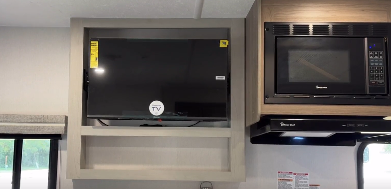 Compact RV entertainment area featuring a mounted Smart TV within a light wood frame, positioned adjacent to a built-in microwave and modern cabinetry, optimizing space and functionality.