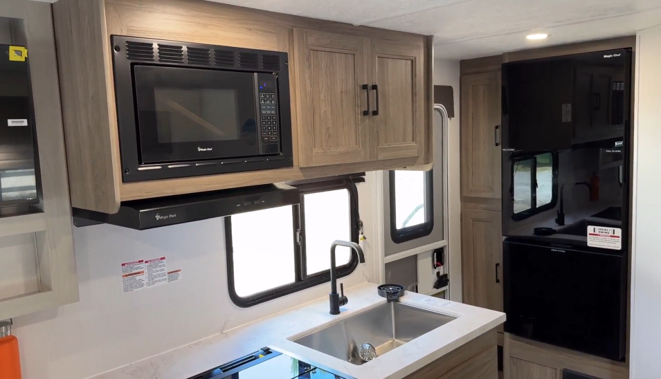 Modern RV kitchen featuring a stainless steel sink with a high-arc faucet, sleek black microwave, ample light wood cabinetry, and a full-size refrigerator, designed for convenience and style in a compact space.