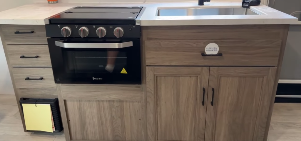 Compact RV kitchen setup featuring a sleek black oven with a modern cooktop, light wood cabinetry with ample storage, and a stainless steel sink with a marble-style countertop, blending functionality and style.