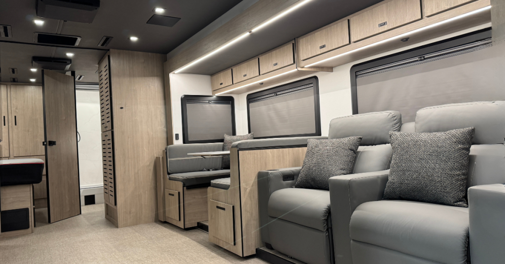 Interior of a modern motorhome showcasing a sleek design with light wood cabinetry, grey recliner seating with throw pillows, and a dining booth with integrated seating. Overhead storage cabinets are illuminated by LED lighting, and large windows with shades provide natural light. A bathroom with a sliding door is visible in the background, contributing to the functional and stylish layout.