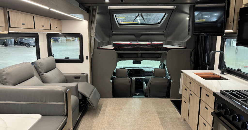 Interior of a motorhome showcasing a well-designed living space with a focus on comfort and functionality. The area includes grey reclining seats with a view of large windows and overhead storage cabinets. The kitchen features light wood cabinetry, a sleek sink, and a stovetop. The cab-over bunk area above the driver’s cockpit is equipped with a skylight, offering additional sleeping space. The seamless transition between the living area, kitchen, and driver’s cab highlights the efficient use of space.