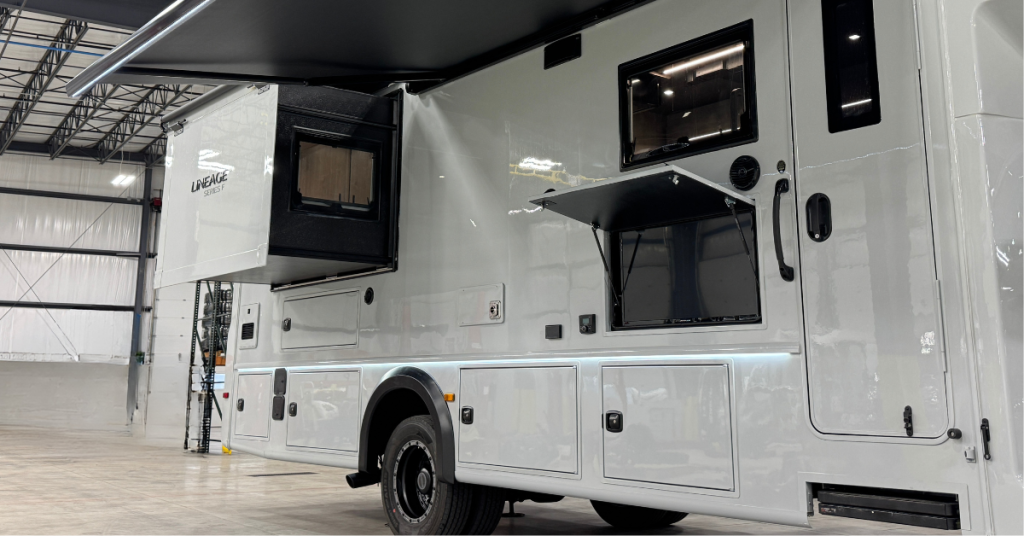 Exterior of a motorhome with a sleek white finish and multiple functional features. The side wall includes storage compartments, a slide-out section for additional interior space, and a fold-up hatch revealing an outdoor entertainment or utility area. A black retractable awning is extended above, providing shade. The motorhome is parked indoors in a spacious facility with industrial lighting.









