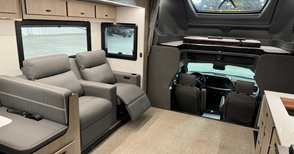 Interior of a motorhome highlighting a cozy and functional living area. The space features two grey recliner chairs with stitching details, overhead storage cabinets, and large windows for natural light. The cab-over bunk area is visible above the driver’s cockpit, equipped with a sleeping platform and windows. The driver's area is accessible via a step-down from the living space, emphasizing seamless design and efficient use of space.