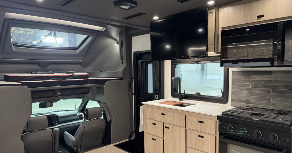 Interior of a motorhome featuring a modern kitchen and entertainment area. The kitchen includes light wood cabinetry, a black stovetop with an oven, a microwave, a deep sink with a black faucet, and a grey backsplash. A mounted flat-screen TV is positioned above the sink. The cab-over bunk area is visible, providing additional sleeping space with a skylight. The driver’s cockpit is located below the bunk, showcasing the efficient use of vertical space.