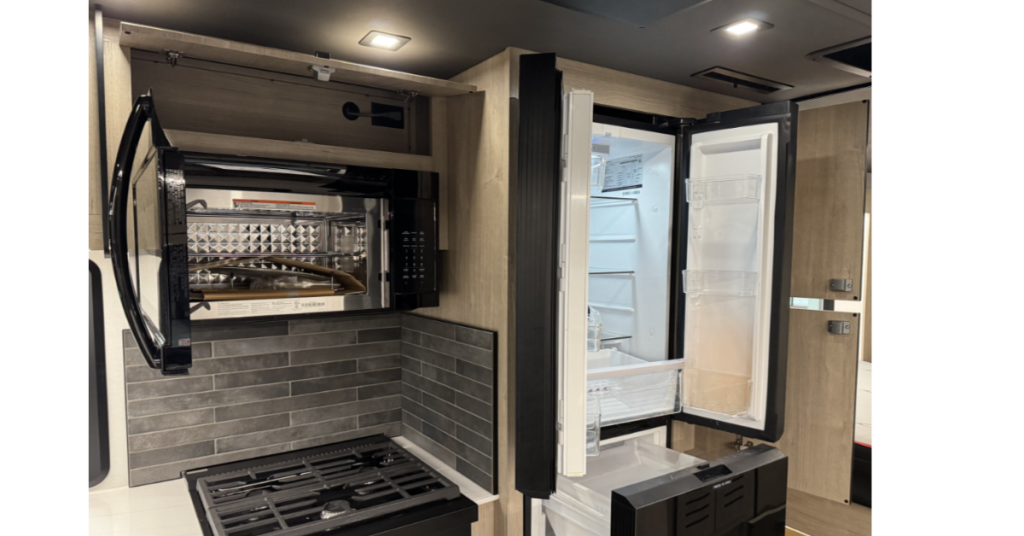 Kitchen area of a motorhome featuring modern appliances and ample storage. A black microwave with a drop-down door is mounted above a grey-tiled backsplash and a three-burner stovetop. Next to it, a sleek, black two-door refrigerator with an open freezer section provides plenty of storage for perishable items. Overhead and adjacent cabinets complement the functional, stylish design.