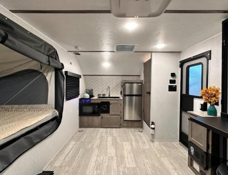 Interior view of a GO Lite travel trailer featuring a cozy sleeping area with a spacious bed dressed in a teal quilt, complemented by a compact fold-out sleeping bunk. The room includes a modern wood-toned nightstand with decorative touches, soft lighting, and a neutral color scheme, creating a relaxing and functional space for campers.