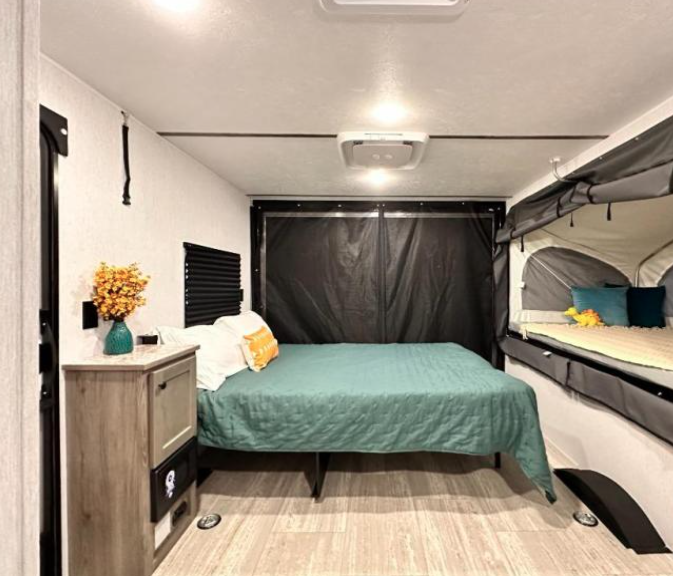 Interior view of a GO Lite travel trailer featuring a cozy sleeping area with a spacious bed dressed in a teal quilt, complemented by a compact fold-out sleeping bunk. The room includes a modern wood-toned nightstand with decorative touches, soft lighting, and a neutral color scheme, creating a relaxing and functional space for campers.