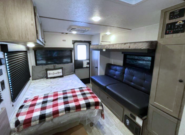 Interior view of a GO Lite travel trailer showcasing a cozy and modern living space. Features include a comfortable fold-down bed with a patterned cover, a leatherette sofa, a top bunk, and wood-toned cabinets for storage. The compact yet functional design includes large windows with blackout shades, efficient lighting, and a durable laminate floor, perfect for on-the-go adventurers.