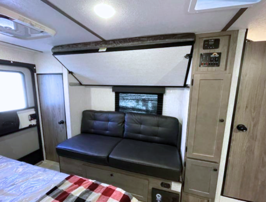 Interior view of a GO Lite travel trailer showcasing a cozy and modern living space. Features include a comfortable fold-down bed with a patterned cover, a leatherette sofa, a top bunk, and wood-toned cabinets for storage. The compact yet functional design includes large windows with blackout shades, efficient lighting, and a durable laminate floor, perfect for on-the-go adventurers.