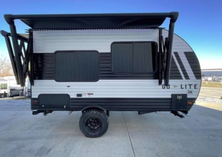 Side view of a GO Lite travel trailer with a fully extended black awning, showcasing its sleek black and white design and off-road tires. The compact and stylish trailer is parked on a paved lot under a clear blue sky, emphasizing its versatility and readiness for outdoor adventures.