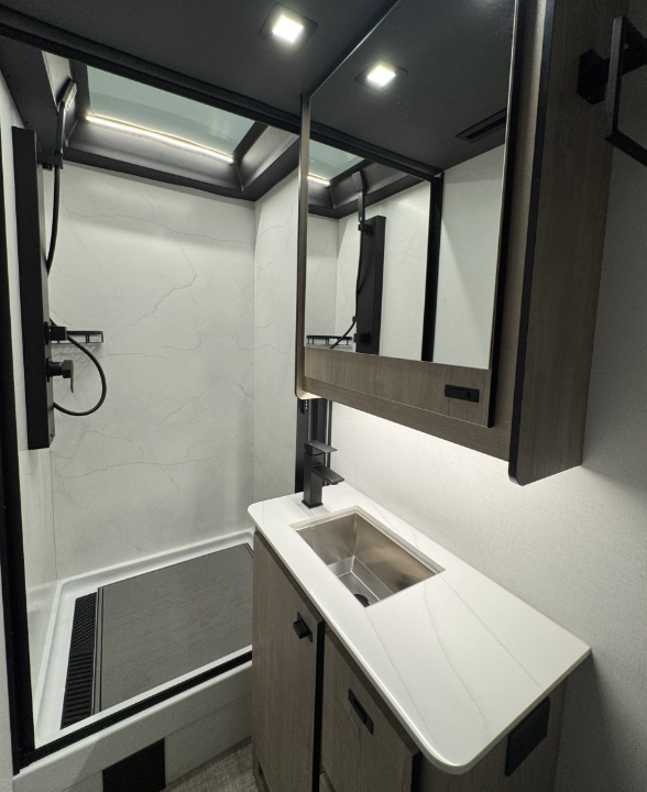 Modern bathroom inside a motorhome featuring a spacious shower with a marble-patterned wall and a black shower fixture. A skylight above the shower provides natural light. The vanity area includes a sleek stainless steel sink with a black faucet, white countertop, and wood cabinetry for storage. A mirrored medicine cabinet adds functionality and enhances the clean, contemporary design.