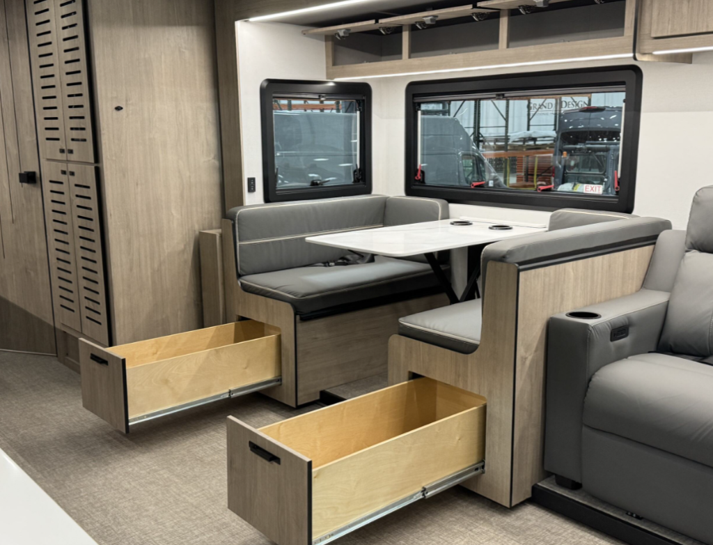 Interior of a motorhome highlighting the dining area with a modern booth-style dinette. The seating includes under-bench pull-out storage drawers, showcasing smart storage solutions. The light wood cabinetry complements the grey upholstery and the marble-like tabletop. Large windows provide natural light, while overhead cabinets offer additional storage, emphasizing functionality and contemporary design.