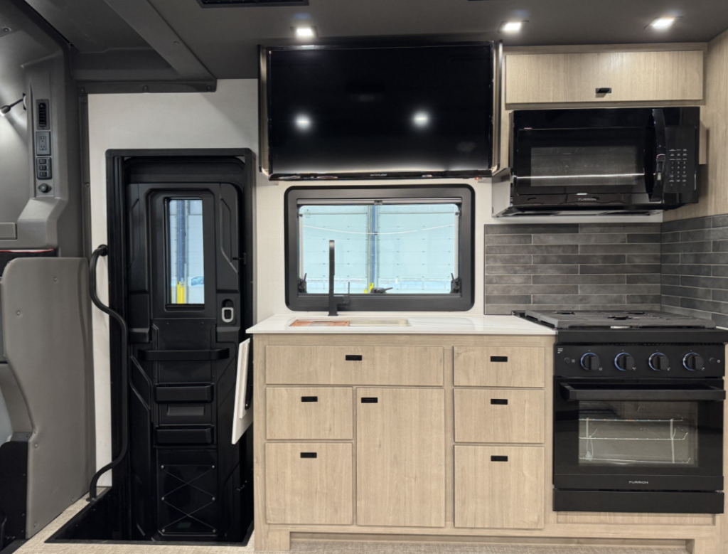 Kitchen and entryway area of a motorhome showcasing a functional and stylish design. The space includes light wood cabinetry, a white countertop with an integrated stainless steel sink and black faucet, and a grey-tiled backsplash. A black three-burner stovetop and oven are paired with a mounted microwave above. A flat-screen TV is installed above the sink for entertainment. To the left, the entry door with a built-in window and handle provides easy access to the motorhome.