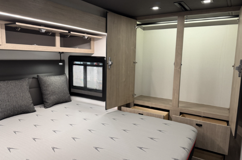 Bedroom area of a motorhome featuring a queen-sized bed with a patterned mattress and grey throw pillows. Overhead storage cabinets with integrated LED lighting provide additional space. A large wardrobe with open doors reveals hanging rods and pull-out drawers for organized storage. A window next to the bed allows natural light, complementing the light wood cabinetry and modern design.