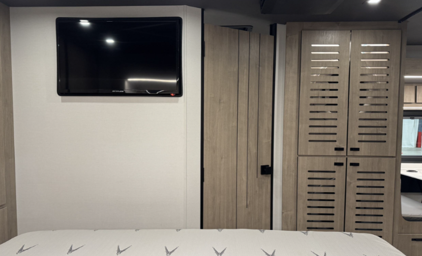 Bedroom area of a motorhome featuring a wall-mounted flat-screen TV for entertainment. The space includes light wood louvered cabinets for ventilation and storage, alongside a folding privacy door that separates the bedroom from other areas. The clean and modern design emphasizes comfort and functionality.