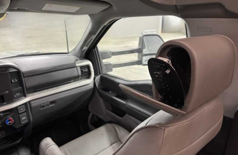 Close-up of a motorhome's front passenger seat featuring a unique headrest storage compartment. The headrest is open to reveal a built-in storage area designed to securely hold electronic devices or small items. The modern dashboard and side mirror are visible in the background, highlighting the vehicle's sleek and practical design.