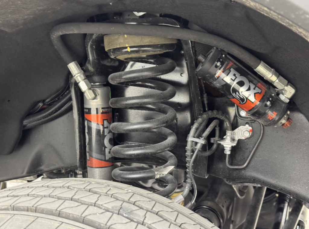 Close-up view of a motorhome's suspension system featuring FOX shock absorbers and coil springs. The robust design highlights advanced suspension technology for improved ride stability and handling. Hydraulic lines and other components are visible, emphasizing the heavy-duty construction suitable for off-road and high-performance driving conditions.