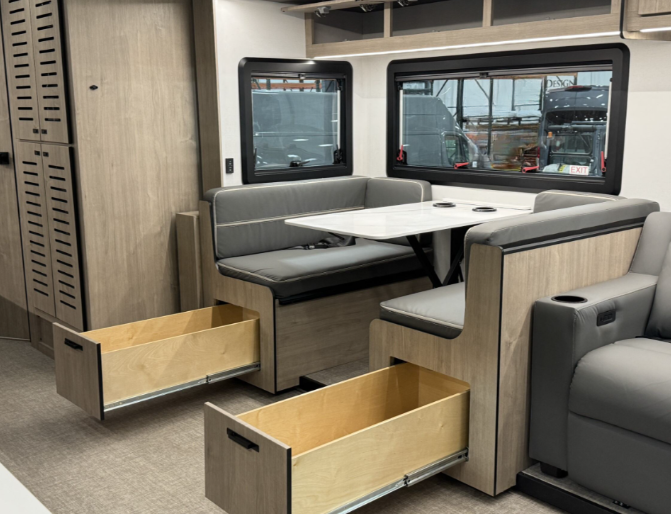 Dining area of a motorhome featuring a booth-style dinette with grey upholstered seating and a white marble-patterned tabletop. The seating includes pull-out storage drawers underneath, providing efficient use of space. Large windows with black frames allow natural light to fill the area, and overhead cabinets offer additional storage. The light wood cabinetry complements the modern and functional design.